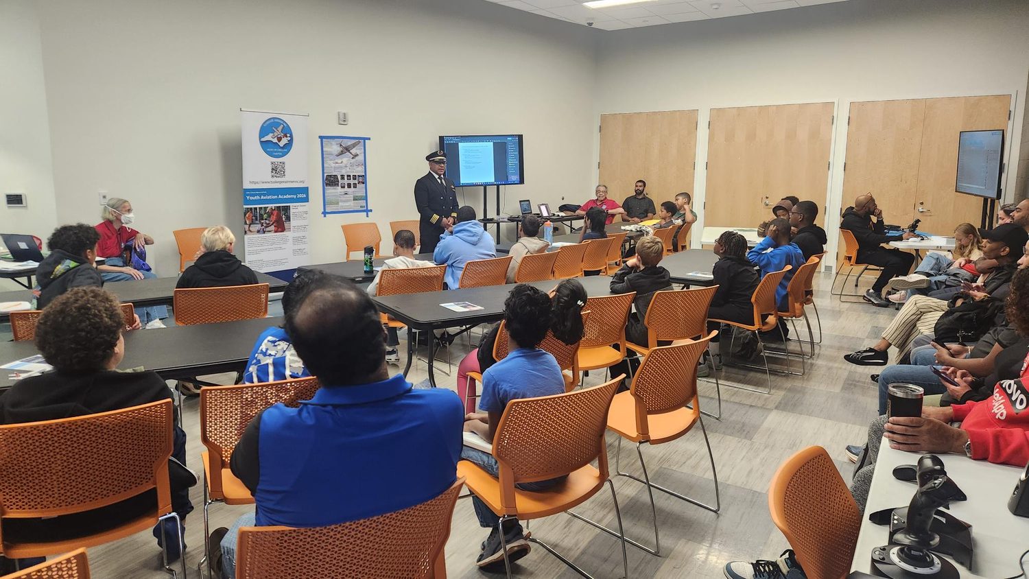 A commercial airline pilot in uniform welcomes students and families at the Youth Aviation Academy, October 2026